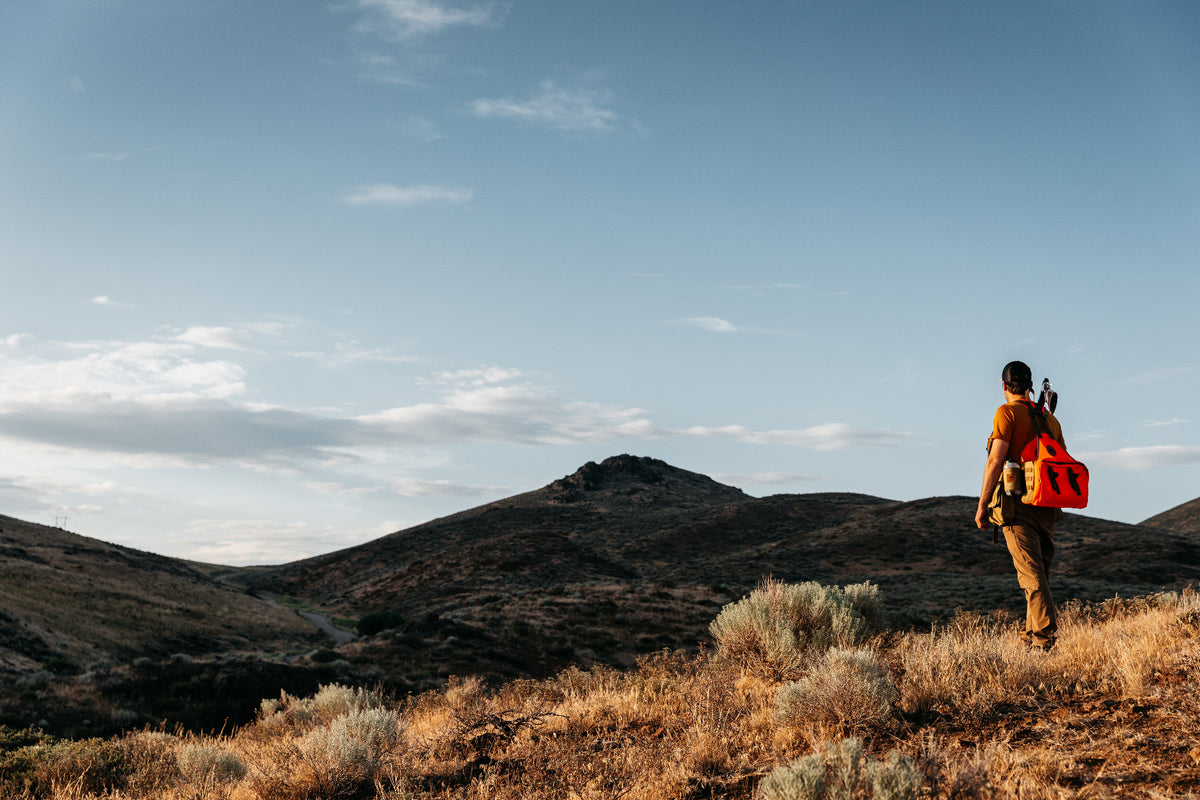 A person hiking through a dry, grassy mountain landscape under a blue sky, wearing an orange Wingworks vest.